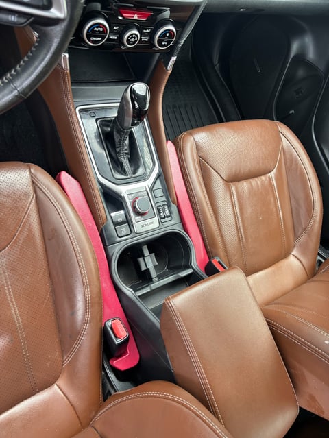 Car interior with tan leather seats, automatic transmission shifter, climate controls, and red seat belt visible